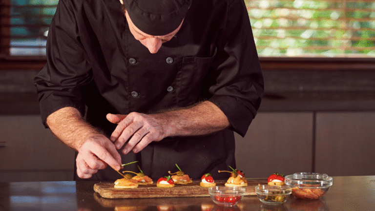Close-up of a professional chef in a black uniform and cap meticulously garnishing gourmet appetizers with chives on a rustic wooden board in a high-end kitchen setting.
