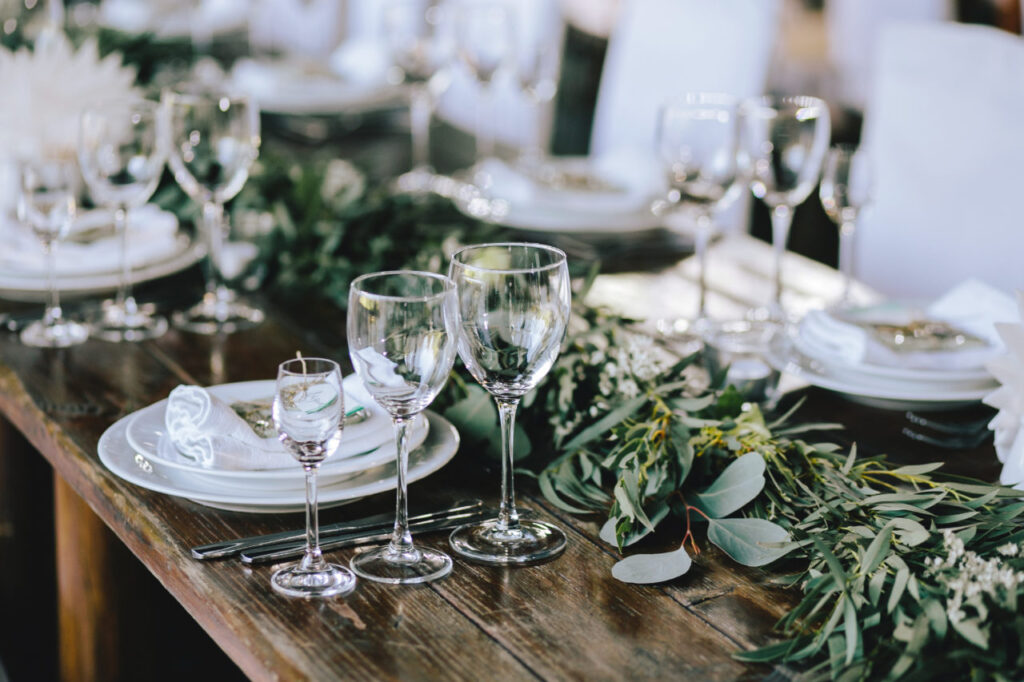 A long wooden dining table elegantly set for a multi-course meal with white plates, crystal wine glasses, white napkins, and a lush green eucalyptus garland centerpiece.