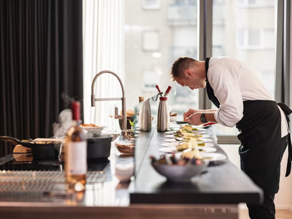 A professional chef in a white uniform and black apron plating a long row of gourmet dishes on a kitchen island for a private event.